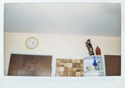 Wide format polaroid photograph of a kitchen wall showing a yellow-rimmed clock, dark wood cabinets, patterned tiles, and a set of small shelves holding a wooden sculpture, an old ketchup bottle labeled as holy water, some crockery and a small blue glass oil lamp.