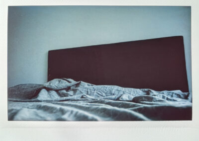 Wide format polaroid photograph of an unmade bed with rumpled blue-grey bedding against a dark grey headboard and pale blue-grey wall.