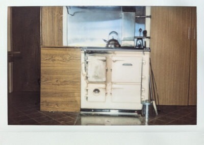 Wide format Polaroid photograph on a small cream coloured range cooker flanked by dark wood kitchen cabinets, with a kettle on top and tiled floor below, in warm vintage tones.