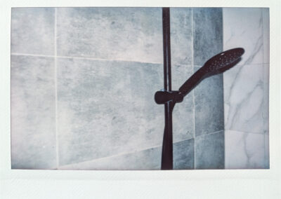 Wide format Polaroid photograph of a dark metal shower head and rail mounted against grey marble-effect bathroom tiles, shot in cool monochrome tones.